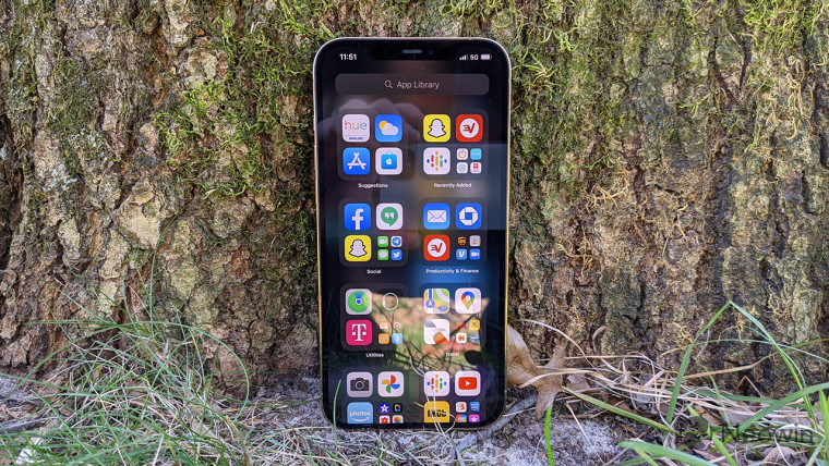 An iPhone 12 Pro Max standing upright on the ground leaning against a tree trunk