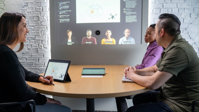 People sitting at a table with a tablet in front and a Teams call with four people on a projector