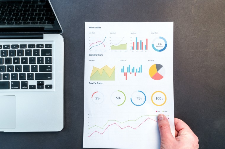 A laptop on the left on a table A persons hand in which a paper containing some data analysis graphs